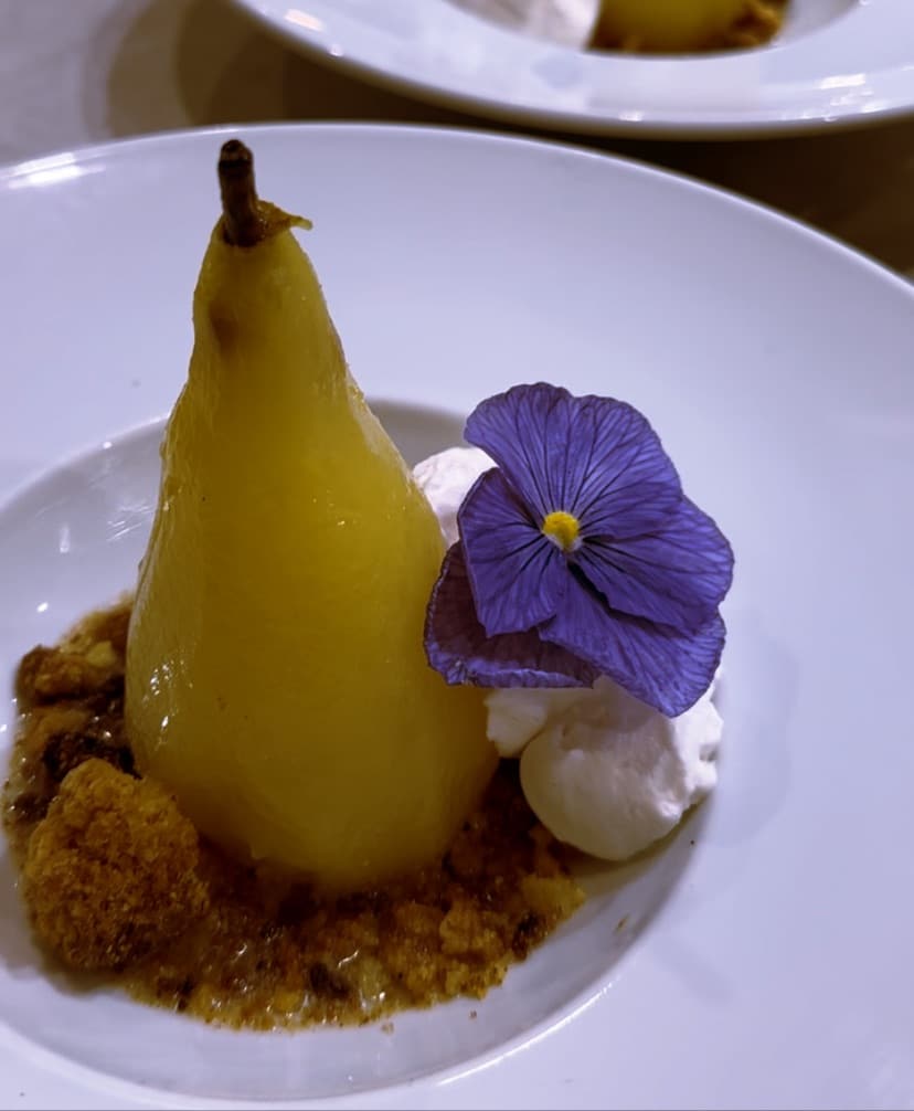 Poire pochée dorée dans une assiette creuse, avec une chantilly, un crumble et une pensée violette