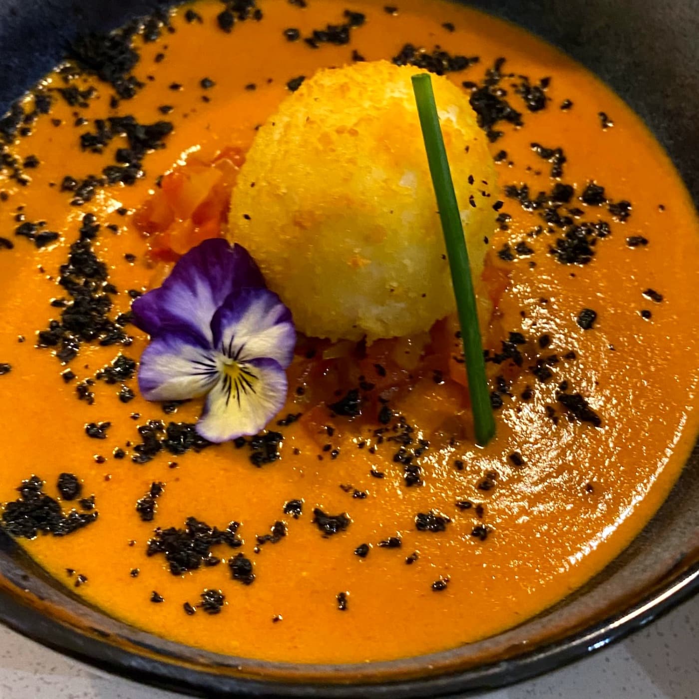 Velouté orange servi dans une assiette creuse sombre, avec une boule dorée, une fleur de pensée et des herbes