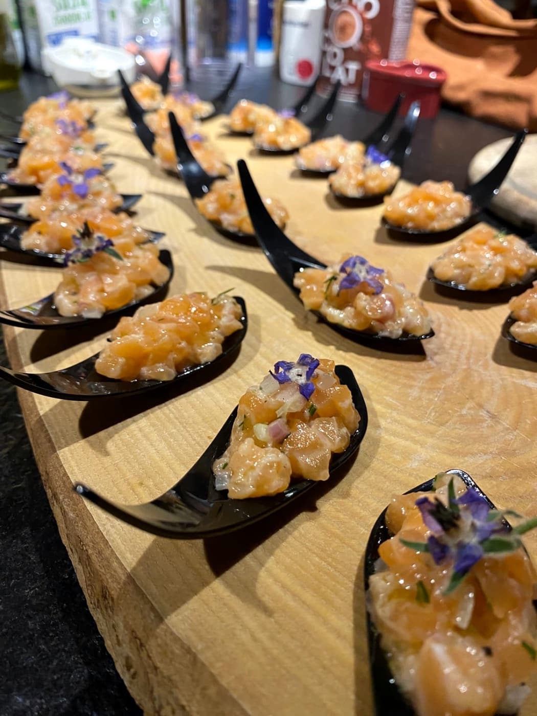 Cuillères noires garnies d’une préparation en dés et de fleurs comestibles, sur une planche en bois