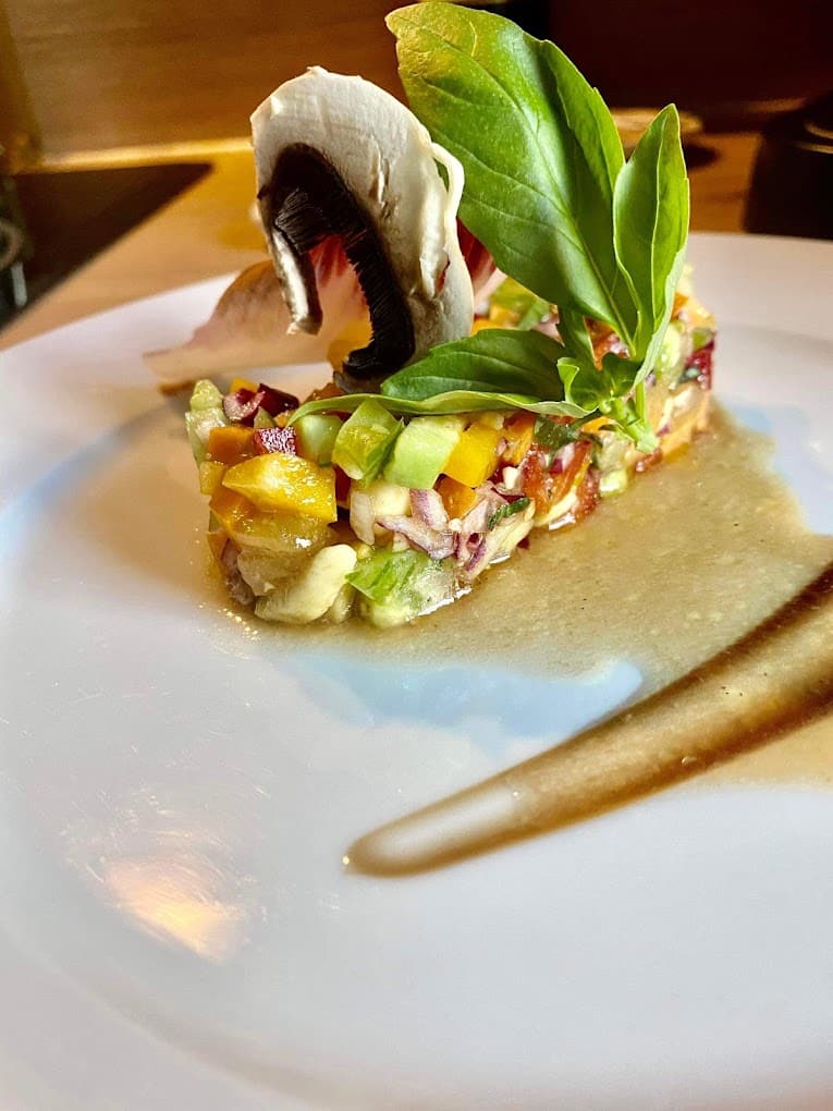 Tartare de légumes colorés dressé sur une assiette blanche, avec une lamelle de champignon, du basilic et une sauce brune